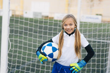 Fröhliches Mädchen in weißer und blauer Uniform mit Fußball, das in die Kamera lächelt, während es allein auf einem grünen Feld in einem modernen Sportverein steht - ADSF08784