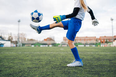 Low angle side view of unrecognizable teenage girl in white and blue uniform and goalkeeper gloves kicking ball while training alone in football arena at stadium in daytime - ADSF08786