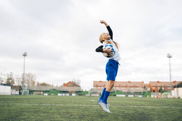 Low angle side view of excited teenage girl in white and blue uniform jumping with arm raised and screaming while celebrating victory in soccer match on green field in contemporary sports club - ADSF08787