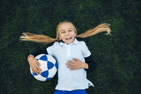 Von oben fröhliches Mädchen mit Pferdeschwanz in Uniform, das lachend mit einem Ball auf einer grünen Wiese im Fußballstadion liegt - ADSF08788