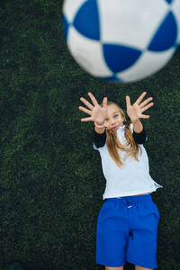 Von oben freudiges Preteen-Mädchen in weißer und blauer Uniform, das einen Fußball wirft, während es allein auf einem grünen Feld in einem modernen Sportverein liegt - ADSF08789