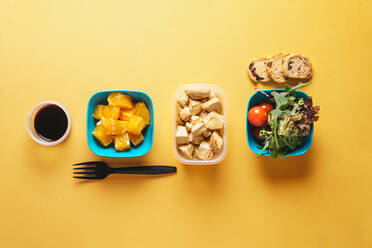 Top view of food containers with bread and tomatoes, arugula and pieces of orange, soy sauce and chicken fillet with black plastic fork on yellow background - ADSF08807