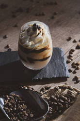 Glas leckeren Sahne-Kaffee-Dessert mit Löffel serviert auf schwarzer Oberfläche mit Kaffeebohnen auf Holztisch - ADSF08829