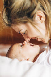 Closeup of glad mom touching nose by nose of baby playing with newborn infant with open mouth having fun lying on bed at home - ADSF08851