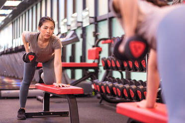 Concentrated young ethnic sportswoman in active wear doing dumbbell row on horizontal bench while training near mirror in modern gym - ADSF08861