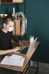 Young man wearing protective mask using laptop on desk at home - ALBF01311