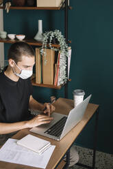 Junger Mann mit Schutzmaske, der zu Hause einen Laptop auf dem Schreibtisch benutzt - ALBF01312