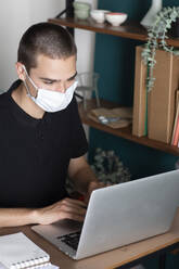 Junger Mann mit Schutzmaske, der zu Hause einen Laptop auf dem Schreibtisch benutzt - ALBF01316