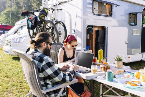 Couple sitting next to camper using laptops on table - DGOF01180