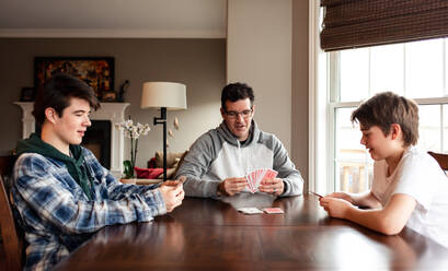 Vater und heranwachsende Söhne spielen gemeinsam Karten am Tisch. - CAVF87917