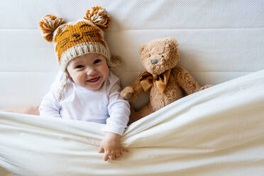 Glücklicher Junge mit Wollmütze liegt mit Teddybär unter der Decke auf dem Bett - CAVF87936