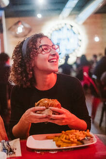 Hungry young female eating tasty burger while sitting at table in brightly illuminated cafe - ADSF08871