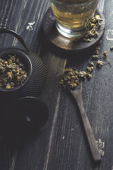 Von oben getrocknetes Gänseblümchen in einem Löffel auf einem dunklen Holztisch neben einer Tasse mit Kräutertee - ADSF08877