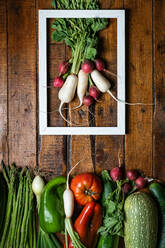 Flat lay of bunch of red and white radish in white frame and set of various fresh vegetables lined below on plank wooden background - ADSF08882