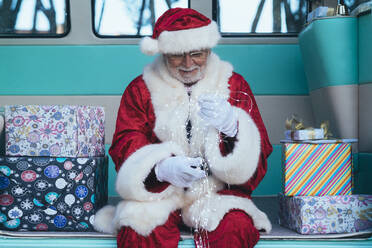 Senior man in costume of Santa Claus sitting in retro van between presents and holding garland in gloved hands - ADSF08894