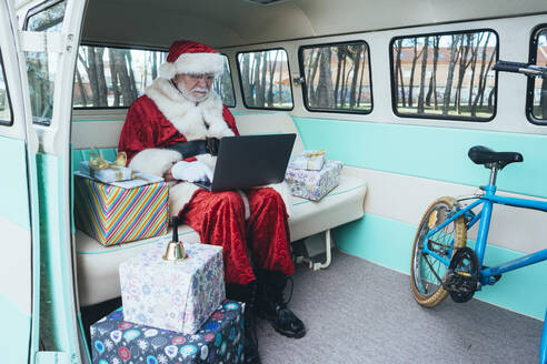 Fröhlicher Mann im Kostüm des Weihnachtsmannes, der mit Laptop und bunten Geschenken an einem sonnigen Tag im Van sitzt - ADSF08896