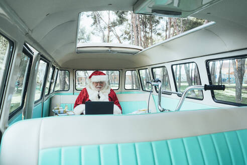 Fröhlicher Mann im Kostüm des Weihnachtsmannes, der mit Laptop und bunten Geschenken an einem sonnigen Tag im Van sitzt - ADSF08897
