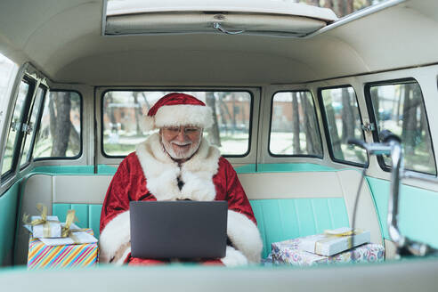 Fröhlicher Mann im Kostüm des Weihnachtsmannes, der mit Laptop und bunten Geschenken an einem sonnigen Tag im Van sitzt - ADSF08898
