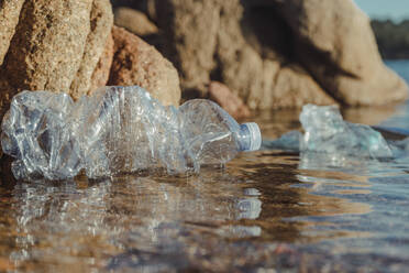 Empty plastic crumpled bottles waste lying in clear water on seaside near rocks - ADSF08903