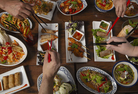 Von oben abgeschnitten unerkennbare Menschen mit Essstäbchen Essen sortiert bunten würzigen thailändischen Essen für Familie Abendessen auf Holztisch serviert - ADSF08924