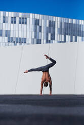 Back view of unrecognizable shirtless sportsman performing handstand with one hand while doing exercises against wall and blue sky on rooftop of modern building - ADSF08932