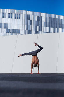 Rückenansicht eines nicht erkennbaren Sportlers ohne Hemd, der mit einer Hand einen Handstand macht, während er auf dem Dach eines modernen Gebäudes Übungen gegen die Wand und den blauen Himmel macht - ADSF08932
