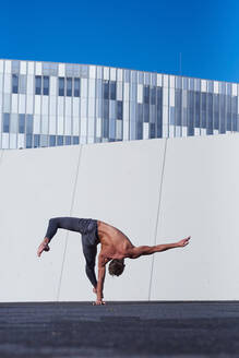 Rückenansicht eines nicht erkennbaren Sportlers ohne Hemd, der mit einer Hand einen Handstand macht, während er auf dem Dach eines modernen Gebäudes Übungen gegen die Wand und den blauen Himmel macht - ADSF08933