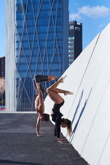 Side view of fit shirtless man athlete and woman performing handstand against wall on rooftop against modern skyscraper on sunny day - ADSF08938