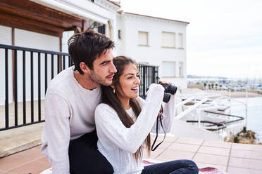 Seitenansicht eines fröhlichen, romantischen Paares in Freizeitkleidung, das mit einem Fernglas auf der Terrasse sitzt und die Aussicht genießt, während es gemeinsam Urlaub am Meer macht - ADSF08940