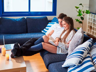 Seitenansicht eines fröhlichen jungen Mannes und einer Frau in Freizeitkleidung, die mit Laptop und Buch auf der Couch sitzen und das Wochenende zu Hause verbringen - ADSF08941