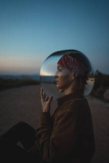 Frau mit einem Fischglas auf dem Kopf, die in der Abenddämmerung auf einer Straße auf dem Lande sitzt - RCPF00287
