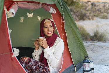 Lächelnde junge Touristin im Hijab in einem Zelt, die einen Becher hält - MPPF00988