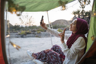 Glückliche junge Touristin mit Hijab in einem Zelt, die ein Selfie macht - MPPF00999