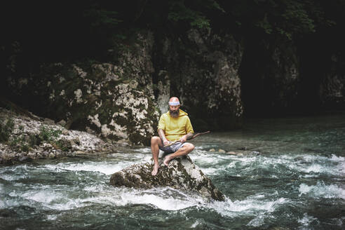 Adventurer with beard sitting in the middle of river on stone and carving on piece of wood - HMEF01044