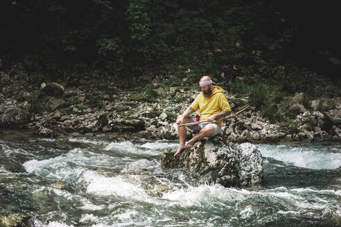 Adventurer with beard sitting in the middle of river on stone and carving on piece of wood - HMEF01045