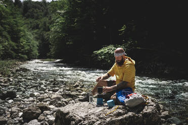 Wanderer mit Vollbart und gelbem Kapuzenpulli bei der Pause, Tee kochen am Flussufer - HMEF01047