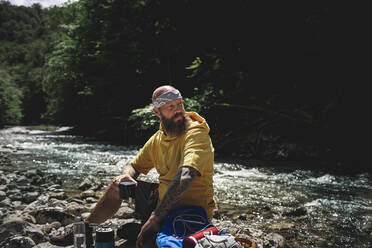 Wanderer mit Vollbart und gelbem Kapuzenpulli bei der Pause, Tee kochen am Flussufer - HMEF01048