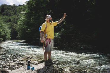 Wanderer mit Vollbart und gelbem Kapuzenpulli steht am Flussufer und benutzt sein Smartphone - HMEF01049