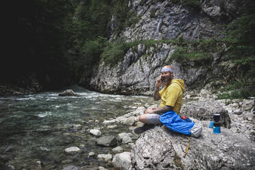 Wanderer mit Vollbart und gelbem Kapuzenpulli sitzt auf einem Stein neben dem Fluss und benutzt sein Smartphone - HMEF01051