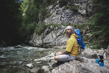 Hiker with full beard and yellow hoodie sitting on stone next to river - HMEF01052