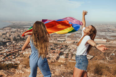 Zwei fröhliche junge Frauen mit einer LGBT-Flagge über der Küstenstadt Almeria, Spanien - MIMFF00131