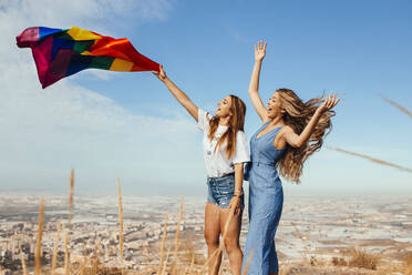 Zwei fröhliche junge Frauen mit einer LGBT-Flagge über der Küstenstadt Almeria, Spanien - MIMFF00132