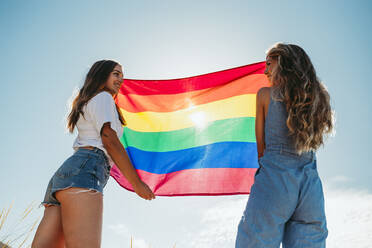 Two smiling young women holding an LGBT flag under blue sky - MIMFF00135