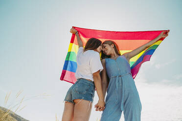 Two affectionate young women holding an LGBT flag under blue sky - MIMFF00136