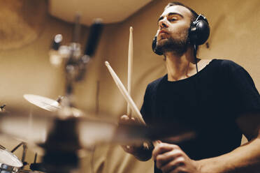 Positive handsome male with headphones playing on drums in studio - ADSF08944