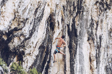 Side view of unrecognizable male in shorts climbing up rough cliff on sunny day in countryside - ADSF08953