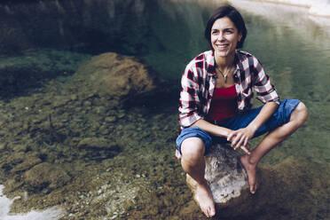 Glückliche erwachsene Frau sitzt auf einem Felsen im ruhigen transparenten Wasser eines Sees und genießt die Natur und lächelt vor sich hin - ADSF08957