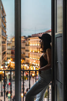 Seitenansicht einer jungen schlanken Frau mit Mütze, die in die Kamera schaut und auf einem Balkon in der Nähe einer beleuchteten Straße am Abend steht - ADSF08977