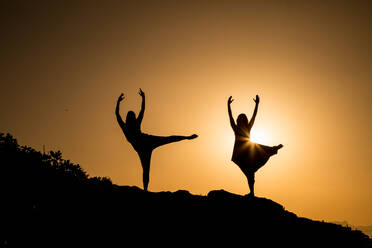 Junge Ballerinas mit gehobenen Beinen und Händen tanzen auf einem Hügel in der Dunkelheit bei Sonnenuntergang - ADSF09060