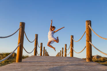 Junge Ballerina in weißer Kleidung mit gehobenem Bein in der Luft auf einem Steg und blauem Himmel an einem sonnigen Tag - ADSF09061